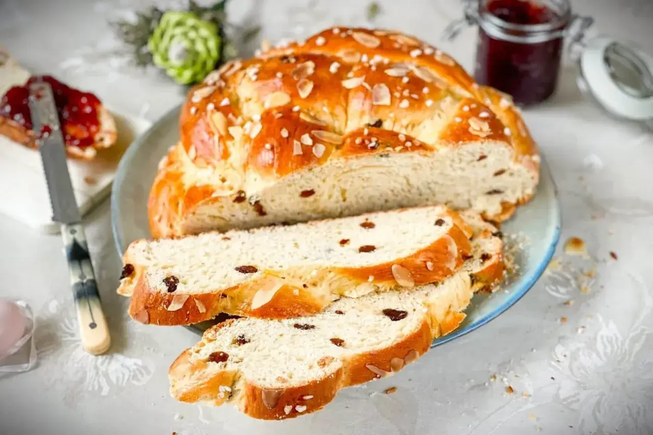 Ein goldbraunes Osterbrot mit Rosinen und Mandelsplittern, perfekt für ein Osterbrot Rezept. Daneben Marmelade und ein Messer.