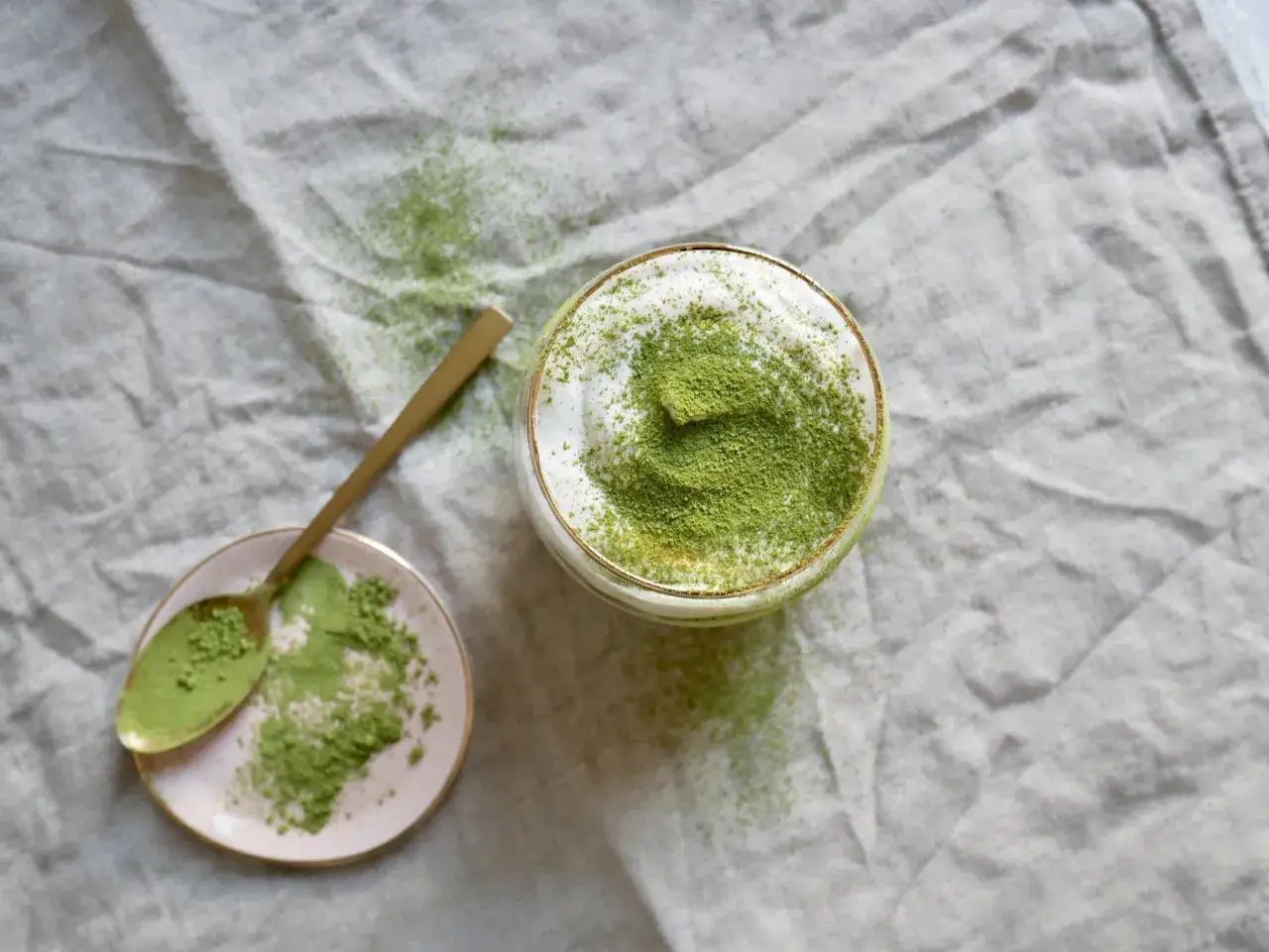 Ein Glas mit cremigem Matcha Tiramisu, best&auml;ubt mit feinem Matcha-Pulver. Daneben ein L&ouml;ffel auf einer kleinen rosa Schale, ebenfalls mit Matcha bestreut.