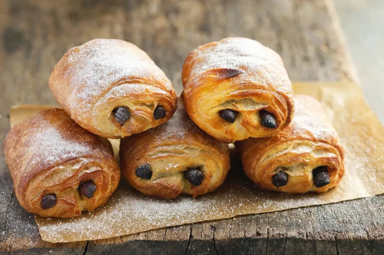 F&uuml;nf goldbraune, mit Puderzucker best&auml;ubte Pain au Chocolat liegen auf Backpapier auf einer Holzoberfl&auml;che.