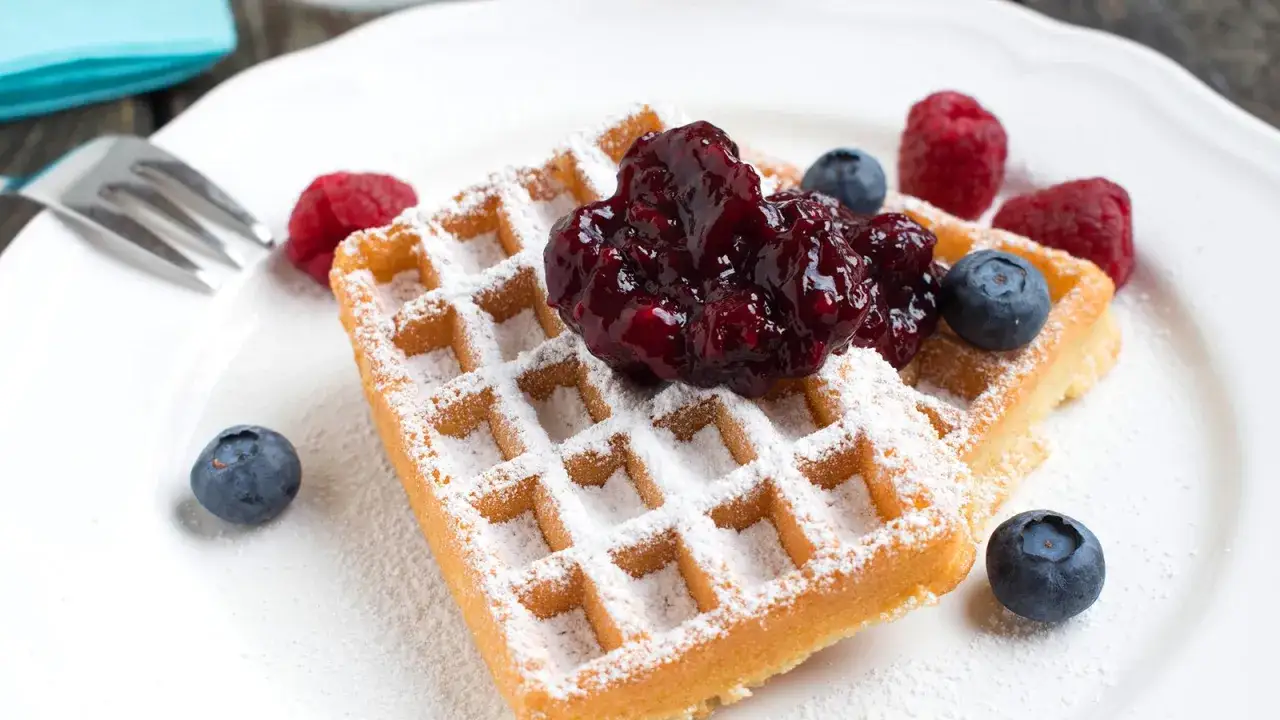 Zwei Buttermilch Waffeln mit Puderzucker, Beeren und Marmelade auf einem wei&szlig;en Teller.
