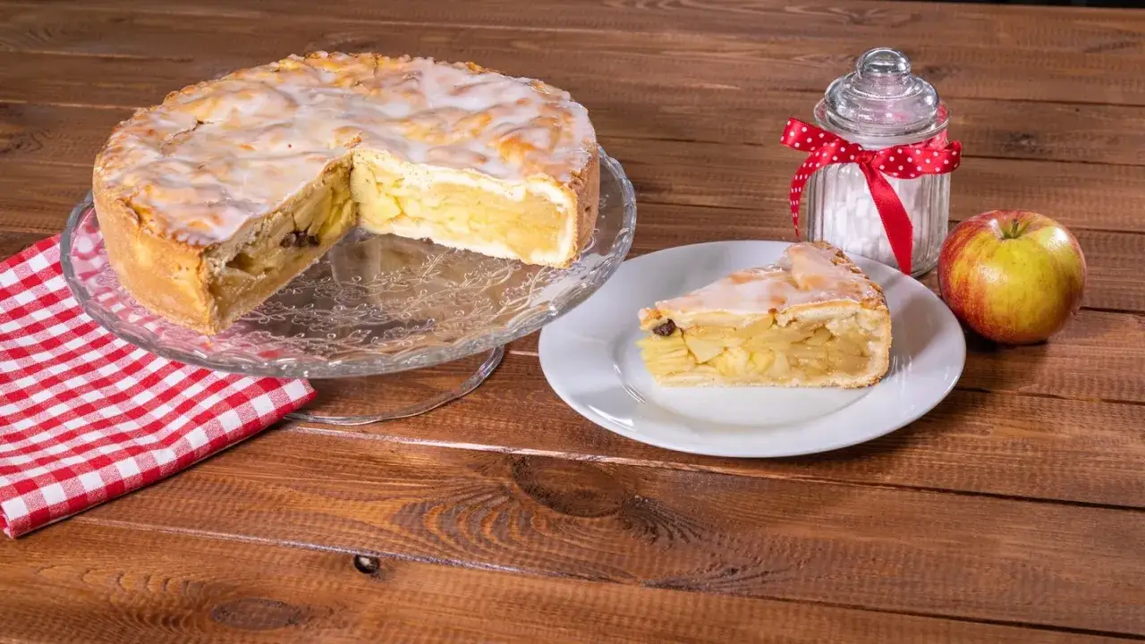 Omas weltbester Apfelkuchen, saftig und mit Zuckerguss überzogen, wartet auf einem Tortenständer. Ein Stück auf einem Teller, daneben ein Apfel und eine Zuckerdose.