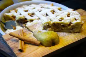 Ein köstlicher Birnenkuchen mit Gittermuster, bestäubt mit Puderzucker. Daneben liegen eine Birne und Zimtstangen – perfekt für ein Birnenkuchen Rezept.