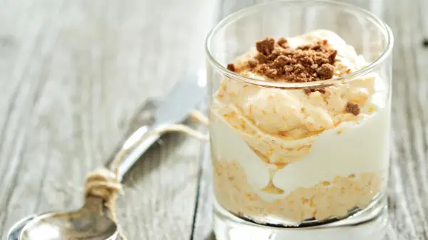 Cremiger cheesecake im glas, geschichtet mit weißer Creme und garniert mit zerbröselten Keksen. Ein Löffel liegt daneben.