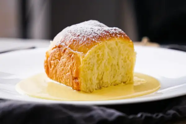 Ein goldbraunes Buchtel, mit Puderzucker bestäubt, ruht in einer cremigen Vanillesauce. Ein köstliches Buchtel Rezept zum Nachbacken.