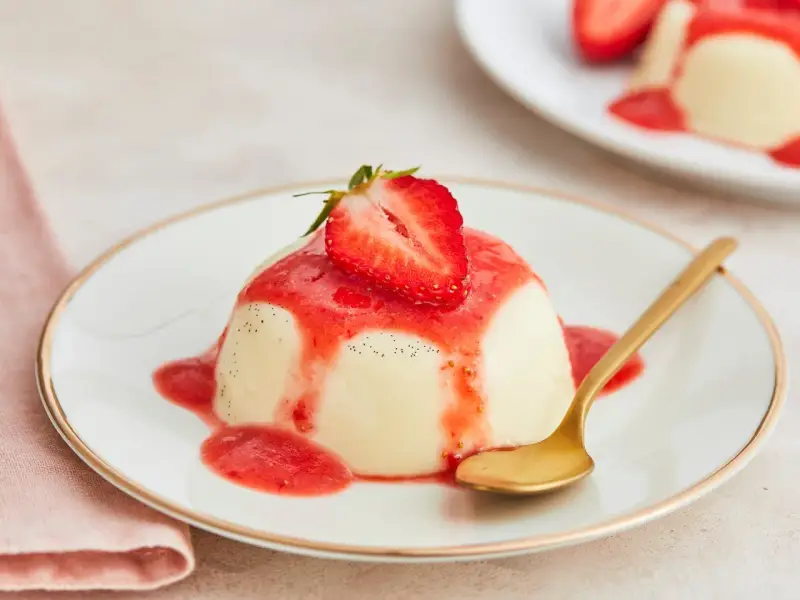 Cremige Panna Cotta mit Erdbeersauce und frischer Erdbeere, garniert mit Vanillepunkten. Eine goldene Löffel liegt daneben.