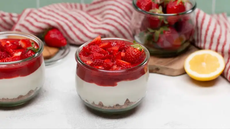 Zwei Gläser mit cremigem Erdbeer-Cheesecake, garniert mit frischen Erdbeeren und Erdbeersauce. Daneben ein Glas voller Erdbeeren und eine Zitronenhälfte.
